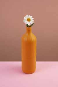 A single white daisy flower with a yellow centre placed inside an orange painted bottle, set on a pink surface against a warm brown background — handmade craft still life photography